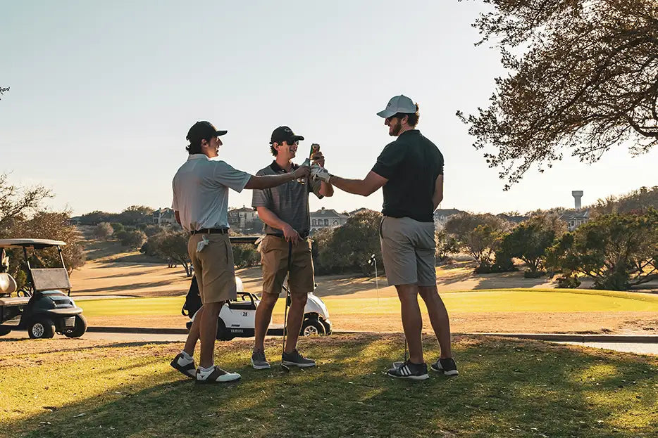 Three golfers clinking drinks together on course at golden hour — social celebration